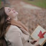 Cheerful young woman with red leaf enjoying life and weather while reading book in autumn park