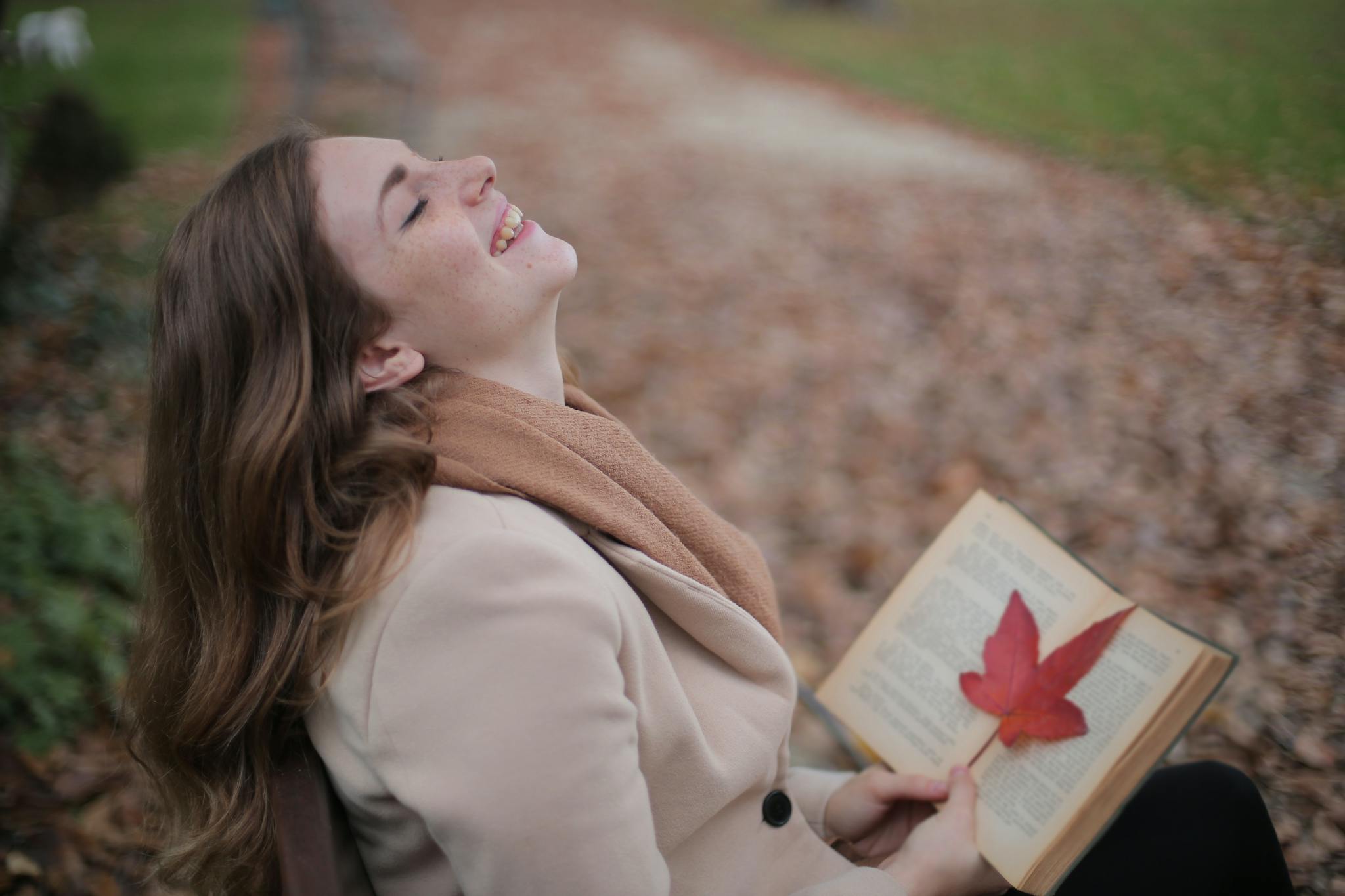 Cheerful young woman with red leaf enjoying life and weather while reading book in autumn park