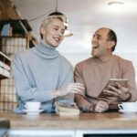 Delighted adult ethnic man laughing and looking at wife while watching together funny video on tablet and drinking coffee in sunny morning at home