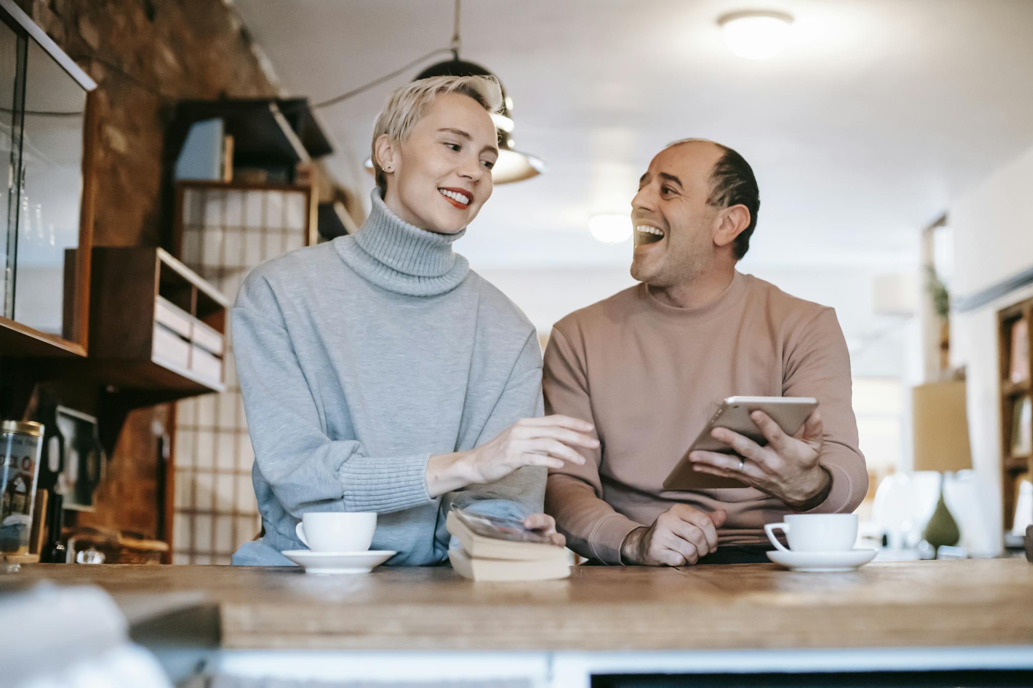 Delighted adult ethnic man laughing and looking at wife while watching together funny video on tablet and drinking coffee in sunny morning at home